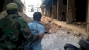 L'image montre une scène de guerre urbaine où deux individus se trouvent dans un environnement dévasté. On peut voir un soldat en uniforme qui tient une personne par l'épaule, tandis qu'un autre homme est visible au loin, apparemment dans une position de menace. Le sol est rempli de décombres, et l'atmosphère générale semble tendue et dangereuse, illustrant les ravages d'un conflit armé. Les bâtiments environnants sont endommagés et abandonnés.