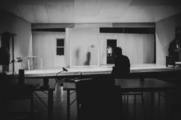 L'image montre un espace scénique avec des murs en bois clair et un éclairage tamisé. On peut apercevoir une silhouette floue à l'arrière-plan, probablement en train de se déplacer. Au premier plan, une personne est assise de dos à une table, observant la scène. L'ambiance est sombre et intimiste, suggérant un moment d'attente ou de concentration avant une représentation. Les éléments de décor, comme les fenêtres et un meuble, ajoutent à l'atmosphère théâtrale.