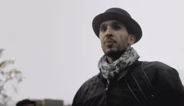 L'image montre un homme portant un chapeau noir et une écharpe. Il a une expression sérieuse et regarde droit devant lui. Le fond semble flou, suggérant une ambiance extérieure peut-être en milieu urbain ou dans un parc. Le temps semble nuageux, ce qui ajoute une atmosphère un peu sombre à la scène.