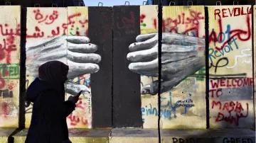 L'image montre un mur avec des graffitis colorés, dont le mot "Révolte" et d'autres messages en arabe et en anglais. Au centre, on peut voir des mains peintes qui semblent se toucher à travers une fissure du mur. Une silhouette d'une personne portant un hijab marche devant ce mur, avec un téléphone à la main. L'ensemble évoque des thèmes de résistance et de lutte sociale.