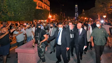 L'image montre une scène nocturne où un groupe de personnes marche dans une rue animée, entouré d'arbres et de lanternes. Au centre, on peut voir des individus en costume, accompagnés par des membres des forces de sécurité, dont l'un est vêtu de vêtements sombres et porte un masque. La foule semble exprimée une certaine agitation, avec des gestes de la main et des expressions variées sur les visages. L'atmosphère générale est tense, et il y a un contraste entre les personnes en tenue formelle et le cadre plus informel de la mobilisation populaire.