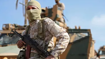 در این تصویر، یک سرباز مسلح با پوشش صورت، در حال ایستادن در پیش‌زمینه قرار دارد. او یونیفورم نظامی و کلاهی به سر دارد و یک اسلحه در دستش است. در پس‌زمینه، یک خودروی نظامی دیده می‌شود و تعدادی از سربازان دیگر در حال فعالیت هستند. آسمان آبی و آب و هوای روشن، صحنه را روشن می‌کند. این تصویر حس قدرت و دقت نظامی را به تصویر می‌کشد.