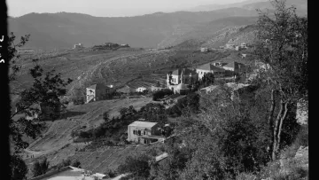 L'image montre un paysage montagneux avec des collines qui s'étendent à l'horizon. On peut apercevoir plusieurs maisons éparpillées sur les flancs des collines, entourées de végétation. La scène semble être prise en noir et blanc, ce qui lui donne un aspect nostalgique. Les terrains agricoles sont visibles, indiquant une zone rurale. Les montagnes en arrière-plan ajoutent une profondeur à la composition paysagère.