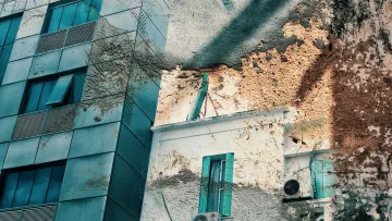 L'image montre un bâtiment moderne avec des façades en verre et en béton, juxtaposed à un mur plus ancien qui semble en bon état. À gauche, les fenêtres et le revêtement du bâtiment présentent des tonalités de bleu et de vert. À droite, on aperçoit une partie du mur texturé, avec des signes d'usure et des éraflures, donnant un contraste intéressant entre le nouveau et l'ancien. On voit aussi des climatiseurs fixés au mur, ainsi que des volets aussi de couleur bleu, ajoutant du caractère à la scène. La lumière joue sur les surfaces, créant un effet visuel captivant.