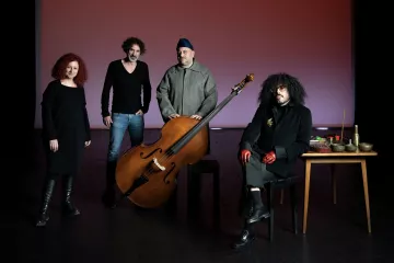 L'image présente un groupe de quatre personnes posant ensemble sur scène. Au centre, il y a un grand instrument de musique, probablement un instrument à cordes, qui attire l'attention. À gauche, une femme avec des cheveux roux bouclés se tient avec une expression sérieuse. À côté, un homme avec des cheveux frisés et un pull noir semble également concentré. Au centre, près de l'instrument, un homme porte une tenue grise et un couvre-chef. Enfin, à droite, un autre homme avec des lunettes et des gants rouges est assis à une table avec divers objets, ayant l'air intrigant. L'éclairage de la scène crée une ambiance artistique et captivante.