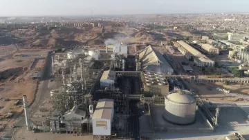 L'image présente une vue aérienne d'un complexe industriel. On peut observer plusieurs bâtiments de grande taille, comprenant des structures cylindriques et pyramidales. La zone environnante semble aride avec des collines et des bâtiments à l'arrière-plan. Des tuyaux et des infrastructures industrielles sont visibles, indiquant une activité de production. On discerne également de la vapeur s'échappant d'une des installations, suggérant un processus en cours. L'ensemble dénote un environnement industriel dans une région désertique.