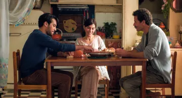 L'image montre trois personnes assises autour d'une table dans une cuisine chaleureuse et accueillante. Il y a un homme à gauche, habillé d'un pull bleu, qui sert de la nourriture à une femme souriante au centre, vêtue d'une blouse claire. À droite, un autre homme, portant un pull gris, semble également engagé dans la conversation. La cuisine est décorée de façon colorée, avec des objets et des ustensiles visibles en arrière-plan, créant une atmosphère conviviale et familiale.