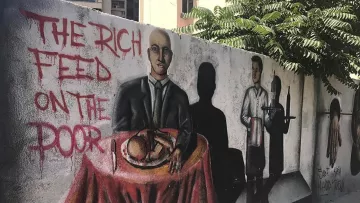 L'image montre une fresque murale colorée sur un mur. Au premier plan, un homme en costume est assis à une table couverte d'une nappe rouge, avec un plat à manger devant lui. À côté, on peut voir une ombre d'un serveur, suggérant un contexte de service alimentaire. Sur le mur, il est écrit "THE RICH FEED ON THE POOR", ce qui évoque un message critique sur les inégalités sociales. Des éléments de l'arrière-plan, comme des bâtiments et de la verdure, ajoutent à l'environnement urbain.
