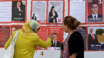 تظهر الصورة جدارًا مغطى بمجموعة من الملصقات الانتخابية. هناك سيدتان تقفان أمام الملصقات. إحداهما ترتدي عباءة ملونة وتظهر وهي تشير إلى أحد الملصقات. يتواجد ملصقات لمرشحين مختلفين، يحمل كل منها صورة واسم المرشح ورقمًا. تعكس الصورة أجواء الانتخابات والحماس المحيط بها، حيث تبدو السيدتان مهتمتين بمناقشة المعلومات المعروضة على الملصقات.
