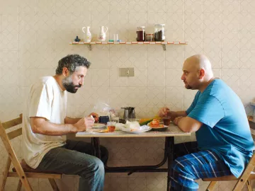 L'image montre deux hommes assis à une table dans une pièce au décor simple. Ils semblent en pleine conversation, chacun portant une expression sérieuse. Sur la table, il y a des assiettes, des verres et des aliments. L'un des hommes porte un t-shirt gris, tandis que l'autre est vêtu d'un t-shirt bleu. La pièce est éclairée de façon naturelle, et le mur est décoré d'un motif en damier. L'ambiance semble à la fois intime et chargée.