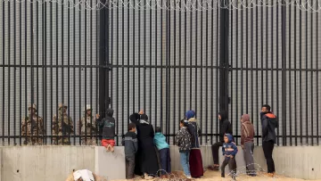 تظهر الصورة مجموعة من الأشخاص الواقفين أمام حواجز حديدية أو سياج مسنن، حيث يواجهون جنود أو عناصر أمنية على الجانب الآخر. يبدو أن هناك أطفالًا ونساء ورجالًا، وبعضهم يحملون أكياسًا أو متعلقات أخرى. البيئة المحيطة موحشة، مع وجود سلك شائك في المقدمة، مما يوضح التوتر والمشاعر القاسية في هذا المشهد.