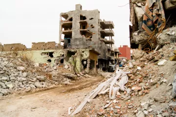 L'image montre un paysage urbain dévasté par la guerre ou une catastrophe. On peut voir des bâtiments partiellement détruits, présentant des murs troués et des structures effondrées. Des débris encombrent le sol, et des morceaux de béton et des matériaux de construction sont éparpillés partout. La rue semble désolée, soulignant l'impact de la destruction sur l'environnement urbain. Les couleurs sont sombres et tristes, renforçant le sentiment de désespoir et de désolation.