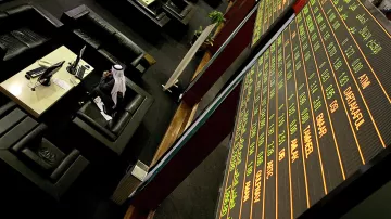 L'image montre un environnement de bourse avec des tables et des sièges confortables. À l'arrière-plan, un grand tableau électronique affiche des informations financières, comme des chiffres et des noms de sociétés. On peut voir quelques personnes, dont un homme en tenue traditionnelle, probablement en train de suivre les fluctuations du marché. L'ambiance semble professionnelle et concentrée, caractéristique d'une salle de négociation.