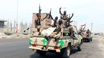 The image depicts a group of armed soldiers riding in the back of a military truck. They appear to be celebrating or expressing excitement, with some of them raising their arms and displaying gestures. The truck has a camouflage pattern, and the background shows a developing urban area, with construction materials and buildings under construction. The overall scene suggests a military convoy or a display of force.