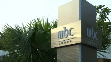تظهر الصورة لافتة تحمل شعار "مجموعة MBC"، وهو معدني وأنيق، ويقع ضمن بيئة نباتية تتضمن أوراق نخيل وشجيرات خضراء. يبدو أن الصورة التقطت في النهار، حيث يظهر ضوء الشمس بشكل جيد. تعكس الألوان والمواد المستخدمة في التصميم الاحترافية والرقي.