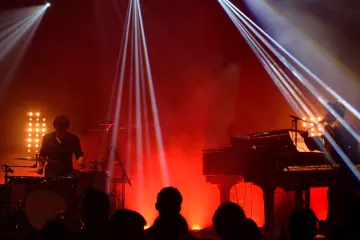 L'image montre une scène de concert avec deux musiciens. À gauche, un batteur joue de la batterie, tandis qu'à droite, un pianiste est assis devant un clavier. L'éclairage est dramatique, avec des faisceaux lumineux qui coupent à travers la fumée ambiante, créant une atmosphère dynamique. En arrière-plan, on devine une audience silhouette, profitant du spectacle. Les couleurs rouges et jaunes ajoutent à l'énergie de la performance.