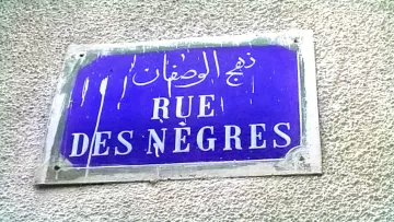 The image shows a street sign. The sign is rectangular and features a blue background with white lettering. It includes text in French that reads "RUE DES NÈGRES," along with Arabic script above it. The sign appears to be weathered, with some signs of wear and tear.