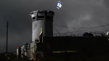 L'image montre une structure semblable à une torre de surveillance, située dans un environnement sombre et nuageux. On peut voir un drapeau israélien flottant au sommet de la tour. Des barbelés sont visibles autour du périmètre, ce qui suggère une zone sécurisée ou protégée. L'éclairage de la scène crée une atmosphère de tension, accentuant l'aspect austère du lieu.