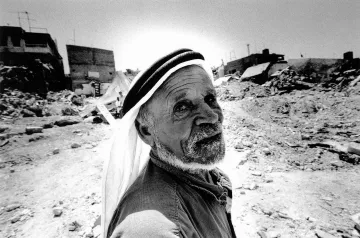 L'image montre un homme âgé au visage marqué, qui regarde directement vers l'objectif. Il porte un keffieh, un couvre-chef traditionnel, et a une expression qui dégage une certaine sagesse ou tristesse. En arrière-plan, on peut apercevoir des ruines, des débris et des bâtiments partiellement détruits, suggérant un environnement difficile ou une situation de conflit. La scène est capturée en noir et blanc, ce qui accentue les textures et les contrastes des éléments environnants.