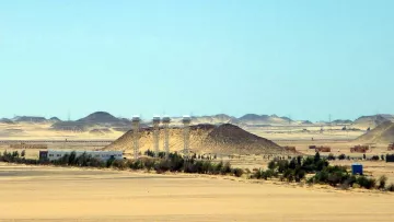 L'image montre un paysage désertique vaste et aride, avec des dunes de sable et des collines. Au centre, on peut distinguer quelques bâtiments qui semblent être des installations humaines, entourés de végétation telle que des arbres ou des buissons. À l'arrière-plan, des collines se découpent contre le ciel, tandis qu'une lumière vive souligne l'immensité du paysage. L'atmosphère semble calme et isolée, typique des régions désertiques.