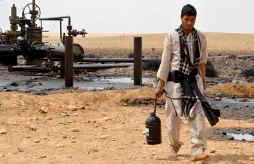 L'image montre un homme marchant à travers un paysage désertique. Il porte des vêtements traditionnels et a un seau noir dans une main. En arrière-plan, on peut voir des installations liées à l'industrie pétrolière, avec des tuyaux et des structures. Le sol est parsemé de pierres et il y a des traces de pétrole sur le terrain. L'environnement semble aride et désertique.