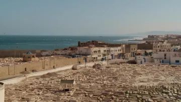 L'image montre un panorama d'une ville côtière, probablement située au bord de la mer Méditerranée. Au premier plan, on peut voir un cimetière avec de nombreuses tombes en pierre, disposées de manière régulière. À l'arrière-plan, il y a des bâtiments blancs, typiques des architectures méditerranéennes, qui bordent la côte. La mer s'étend à l'horizon, avec des vagues visibles, et il semble y avoir une légère brume sur l'eau. La scène dégage une ambiance paisible et sereine, avec une lumière douce évoquant l'heure du soir ou du matin.