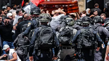 تظهر الصورة مشهدًا من حدث عام حيث يحدث تداخل بين مجموعة من الناس وقوات الأمن. يتم حمل تابوت خشبي من قبل المشاركين في الهتافات، بينما يحيط بهم رجال شرطة يرتدون تجهيزات خاصة. الخلفية تضم أعلامًا، مما يشير إلى أن هذه الفعالية قد تكون مرتبطة بمسألة وطنية أو سياسية. الجو يبدو مشحونًا بالانفعالات، مع وجود حشد من الناس في حالة من التوتر.