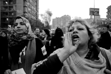 L'image montre une scène de manifestation en noir et blanc. Au premier plan, une femme au cheveux blonds, visiblement passionnée, crie ou exprime une émotion forte, tandis qu'une autre femme, à gauche, porte un foulard et semble attentive. On aperçoit en arrière-plan un grand groupe de personnes, ce qui suggère un mouvement collectif. L'atmosphère est chargée d'énergie et de détermination. Les bâtiments autour ajoutent un contexte urbain à cette démonstration.