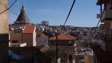 L'immagine mostra un paesaggio urbano con diverse abitazioni disposte su una collina. In primo piano, ci sono edifici con tetti a spiovente di colore rosso e strutture in muratura. Sullo sfondo si riconosce un grande edificio con una copertura a cupola, che si erge tra le case. Il cielo è sereno e limpido, contribuendo a dare un'impressione di vivacità al panorama cittadino. Inoltre, alcuni fili elettrici attraversano l'immagine, evidenziando l'aspetto urbano della scena.