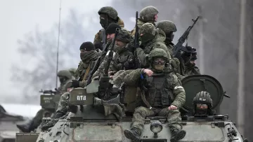 L'immagine mostra un gruppo di soldati equipaggiati con abbigliamento militare e armi, che si trovano su un veicolo blindato. Alcuni di loro indossano maschere o occhiali protettivi, mentre altri appaiono attenti e pronti all'azione. Il contesto è probabilmente un'area di conflitto, con un paesaggio grigio e invernale sullo sfondo. L'atmosfera è seria e tesa, riflettendo la situazione militare.