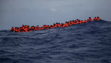 L'image montre un groupe de personnes en mer, portant des gilets de sauvetage orange. Elles semblent rassemblées sur une petite embarcation, luttant contre des vagues dans une étendue d'eau bleue profonde. Le ciel est nuageux, ce qui accentue la gravité de la situation maritime. Cette scène évoque des thèmes de migration, de survie et de danger en mer.