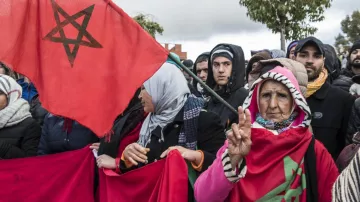 L'image montre une foule rassemblée, avec des gens portant des drapeaux marocains. Parmi eux, on peut voir une femme âgée qui fait un signe de paix avec sa main. Les participants semblent engagés et soutiennent une cause commune, probablement en lien avec leur identité ou des revendications sociales. Leur expression et leur posture suggèrent une forte détermination et un esprit de solidarité. Il y a une atmosphère de mobilisation collective.