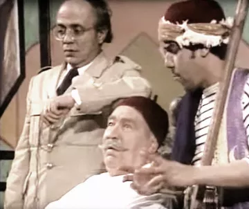 L'image montre trois personnages dans un cadre qui semble être une scène de théâtre ou une comédie. L'un des personnages est assis, portant une blouse blanche et un fez, affichant une expression détendue. À sa gauche se trouve un personnage en tenue militaire, regardant sa montre avec une expression préoccupée. À droite, un troisième personnage, également en fez, joue d'un instrument de musique tout en gesticulant, ajoutant une ambiance dynamique à la scène. L'ensemble évoque un moment comique ou dramatique.