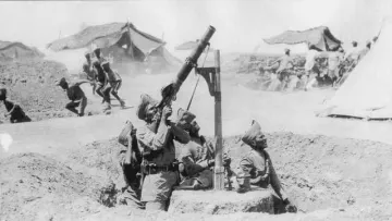 The image depicts a group of soldiers operating a mortar in a wartime setting. They are wearing military uniforms and are positioned in a trench-like area. One soldier is aiming the mortar, while other soldiers are present nearby, possibly providing support or preparing for an attack. In the background, there are tents suggesting a military camp, and additional soldiers can be seen, likely engaged in activity related to the ongoing conflict. The photo has a historical, black-and-white appearance, conveying a sense of the era in which it was taken.