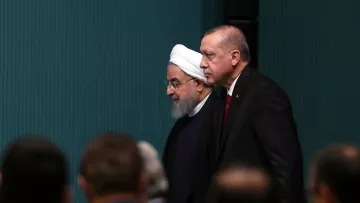 The image shows two men walking side by side. One is wearing a white turban and a black robe, while the other is dressed in a formal suit. They appear to be in a formal setting, likely a conference or meeting, with other individuals partially visible in the foreground. The background features a minimalist wall with a light blue-green color. The atmosphere seems serious and professional.