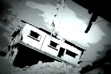 L'image montre un bâtiment en ruine, partiellement détruit, avec des murs endommagés et une atmosphère sombre. La structure semble en déséquilibre, suspendue ou penchée, ce qui renforce l'impression de dévastation. Le ciel est nuageux, ajoutant une tonalité dramatique à la scène. Les teintes sont principalement en noir et blanc, créant un effet visuel frappant.
