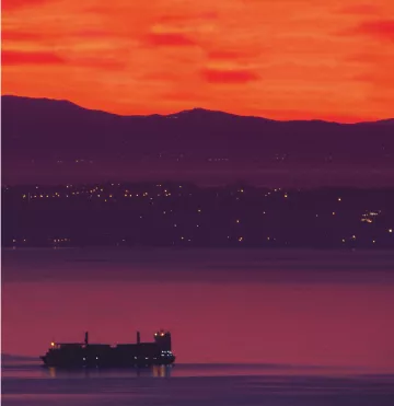 L'image présente un coucher de soleil spectaculaire, où le ciel est teinté de rouge et d'orange lumineux. En arrière-plan, on aperçoit des collines sombres, tandis qu'à l'avant-plan, un cargo navigue sur l'eau calme. Les lumières d'une ville ou d'un port scintillent le long de la côte, ajoutant une touche de vie à cette scène paisible.