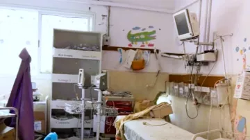 L'immagine mostra una stanza d'ospedale in uno stato di trascuratezza. Si possono notare letti ospedalieri con lenzuola disordinate, attrezzature mediche sparse e un mobile con vari oggetti all'interno. Le pareti sono decorate con disegni infantili, ma l'ambiente appare comunque piuttosto disordinato. Un monitor è appeso al muro e ci sono altri strumenti medici, indicando che si tratta di un'area destinata alla cura dei pazienti. La luce naturale entra dalla finestra, creando un contrasto con l'aspetto poco curato della stanza.