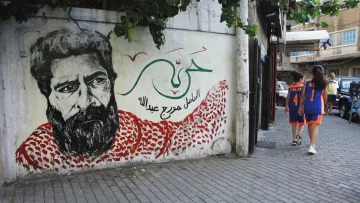 L'image montre une fresque murale d'un homme avec une barbe, probablement un portrait stylisé. Il porte un keffieh rouge et blanc, et le fond est orné de motifs et de couleurs vives. À côté, deux personnes marchent sur un trottoir, portant des vêtements de sport. L'environnement semble urbain, avec des bâtiments en arrière-plan et de la végétation. Les éléments de la fresque et la scène de rue créent une atmosphère vivante et culturelle.