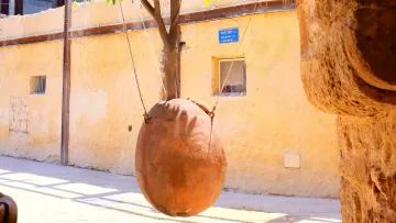 L'immagine mostra un quartiere caratteristico con un muro di colore giallo chiaro. Al centro, si vede un grande vaso di terra cotta che oscilla, sospeso con delle corde. Il vaso sembra appeso a un albero, mentre sullo sfondo si intravede un edificio con strutture in legno e finestre. La scena appare luminosa e vivace, con una sensazione di atmosfera calda e accogliente.