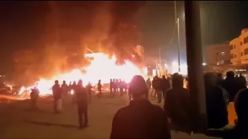در این تصویر، ظاهراً افرادی در یک صحنه شبانه به سوی یک آتش بزرگ در حال تماشای آن هستند. شعله‌های آتش به شدت در حال سوزاندن یک ساختمان یا ساختار دیگر هستند و دود سیاهی به سمت آسمان بلند می‌شود. مردم در گروه‌های مختلف در پس‌زمینه ایستاده‌اند و به سمت محل آتش نگاه می‌کنند. احتمالاً این صحنه به یک حادثه یا اعتراض مربوط می‌شود. روشنایی آتش فضای اطراف را درخشان کرده است.