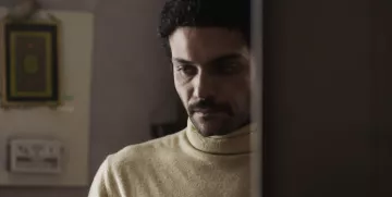 L'image montre un homme avec des cheveux bouclés et une moustache, portant un pull à col roulé de couleur claire. Il semble pensif, regardant vers le bas, avec une expression de réflexion ou de sérieux. En arrière-plan, on aperçoit des éléments de décoration intérieure, mais les détails sont flous. L'atmosphère de l'image dégage une certaine intimité ou mélancolie.
