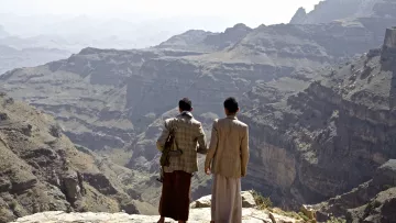 L'immagine mostra due uomini che si trovano su un bordo roccioso, ammirando un vasto paesaggio montano. Le loro spalle sono visibili mentre guardano verso il fondo di una valle profonda e frastagliata. Le montagne circostanti sono di diverse tonalità di grigio e marrone, con una vegetazione scarsa visibile in alcune zone. L'atmosfera appare serena e suggestiva, con un cielo chiaro sullo sfondo.