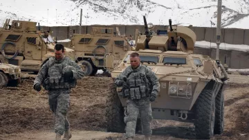 En la imagen se pueden ver a dos soldados caminando por un terreno irregular y arenoso. Están vestidos con uniformes militares y parecen estar en un entorno montañoso, con algunas unidades de vehículos blindados al fondo. La atmósfera es de actividad militar, con un paisaje que sugiere que están en una misión en un área remota. La nieve se puede observar en las montañas que rodean la escena.