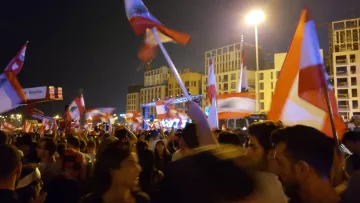 L'image montre une foule rassemblée à la nuit tombée, tenant des drapeaux libanais. Les gens semblent être en plein mouvement, probablement en train de célébrer ou de manifester. On aperçoit une scène à l'arrière-plan, éclairée par des lumières, ce qui suggère un événement festif ou politique. L'atmosphère est dynamique, remplie d'énergie, avec des expressions de joie et d'engagement parmi les participants.