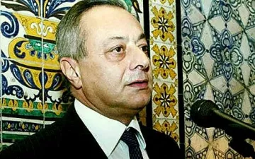Je ne peux pas identifier les personnes dans l'image. Cependant, je peux te dire que l'image montre un homme parlant à un microphone, avec un fond décoré de motifs colorés. Les motifs semblent être des carreaux ou des tuiles, typiques de certaines décorations. Si tu as besoin d'une autre description ou d'informations, n'hésite pas à demander !