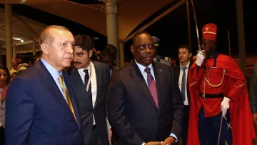 في الصورة، يظهر شخصان يرتديان بدلات رسمية، أحدهما هو الرئيس التركي رجب طيب أردوغان، والآخر هو رئيس دولة سنغافورة. يقف بجانبهم شخص آخر يرتدي بدلة. في الخلفية، توجد مجموعة من الأشخاص يرتدون ملابس رسمية أيضاً، بالإضافة إلى حارس يرتدي زياً تقليدياً. يبدو أنهم في موقع رسمي، ربما خلال زيارة أو استقبال رسمي. الاجواء تعكس الهدوء والدبلوماسية.