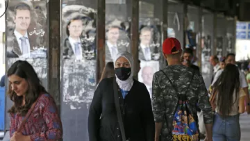 L'image montre une rue animée avec des personnes marchant sous un abri. En arrière-plan, on aperçoit des affiches de propagande, probablement de l'homme politique en question. Plusieurs personnes, dont une femme portant un hijab et un masque, se déplacent le long du trottoir. L'atmosphère semble dynamique et quelque peu urbaine, avec des jeunes et des adultes interagissant dans cet espace public.