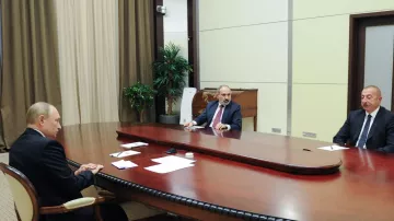 L'image montre une réunion formelle dans une salle ornée. Trois hommes sont assis autour d'une grande table ovale en bois. Au centre, un homme en costume parlant, probablement un chef d'État ou un dirigeant. À sa gauche, un homme au visage sérieux, également en costume. À droite, un autre homme, avec une expression attentive. La pièce est décorée de rideaux et d'une plante d'intérieur, ce qui lui donne un aspect sobre et professionnel.