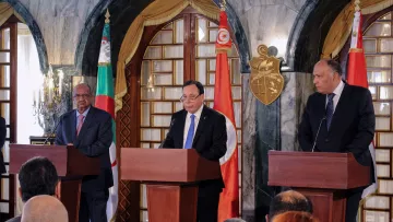 L'image montre une conférence de presse avec trois hommes debout derrière des pupitres en bois. Au centre, un homme prend la parole, tandis que les deux autres se tiennent à sa gauche et à sa droite. Les drapeaux de l'Algérie et de la Tunisie sont visibles en arrière-plan, ce qui suggère que cet événement pourrait concerner une coopération entre ces deux pays. L'ambiance semble formelle et sérieuse, avec un public assis dans la partie inférieure de l'image.